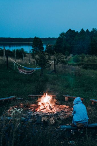 A person with a hood sitting next to the bonfire with hammocks, forest and lake in the background.