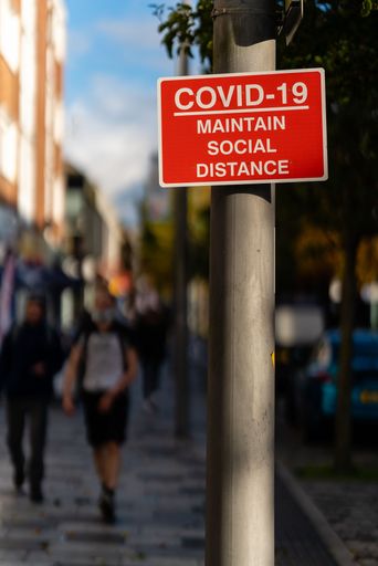 A vertical shot of red and white "COVID-19 Maintain Social Distance" sign on lamp post with pedestrians in the blurred background