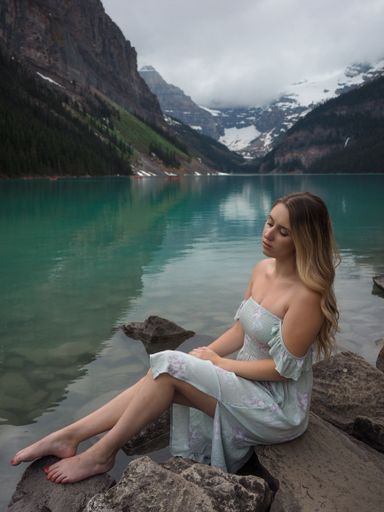 A vertical shot of a beautiful Caucasian girl in a blue dress sitting on the Lake Louise shore