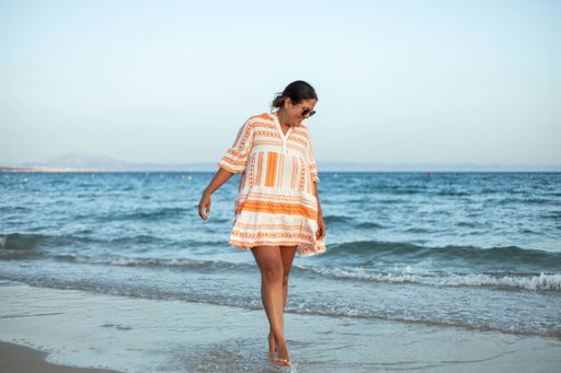 A beautiful Hispanic woman in sunglasses smiling at a sandy beach with a sea in the background