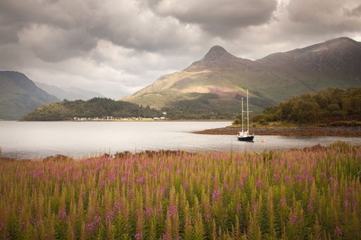 A boat sailing in a lake against a green field and mountains on a cloudy day in Glencoe, Scotland