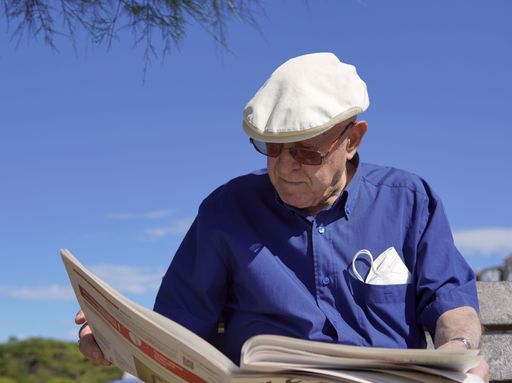 An older Caucasian man reading a newspaper sitting on a park bench