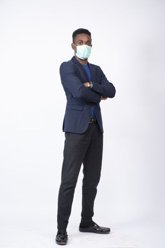 A vertical shot of a young black man wearing a suit and face mask standing confidently with his arms crossed