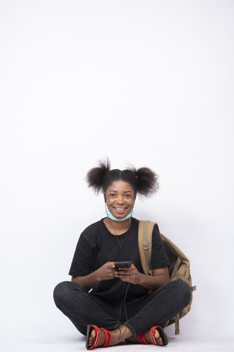 A vertical shot of a pretty young African girl sitting cross-legged with a mask on her jaw and using her mobile phone, smiling