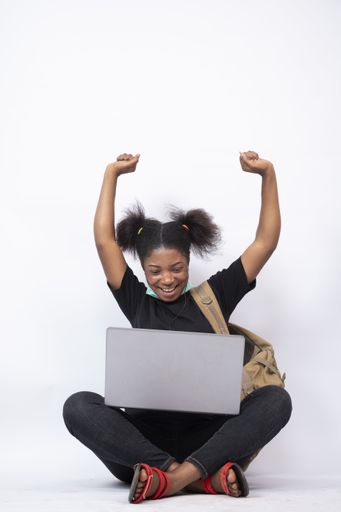 A vertical shot of a beautiful young black woman sitting cross-legged with a mask on her jaw, using her laptop, feeling happy and celebrating