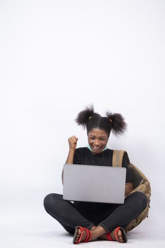 A vertical shot of a beautiful young black woman sitting cross-legged with a mask on her jaw, using her laptop, feeling happy and celebrating