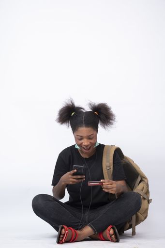 A vertical shot of a young African girl sitting cross-legged, with a mask on her jaw, using her mobile phone and credit card, smiling