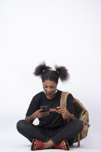 A beautiful young African female carrying a backpack, sitting legs crossed using her mobile phone and credit card