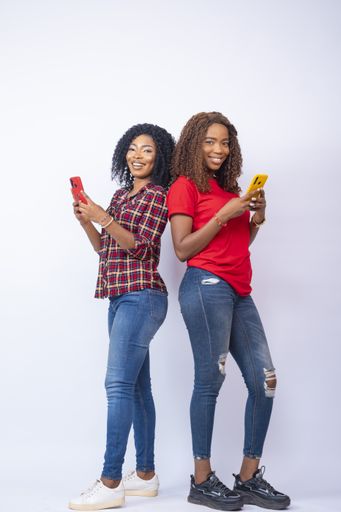 A vertical shot of happy young African women using their mobile phones and standing back to back
