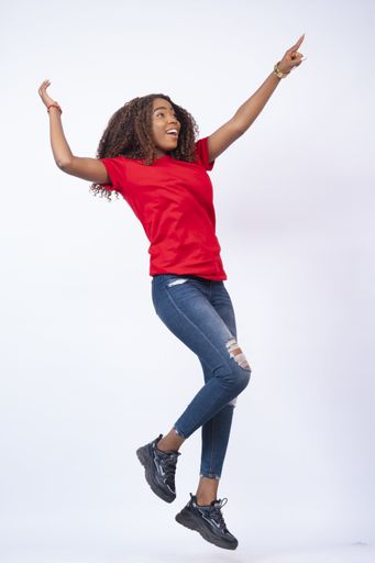 A vertical shot of a pretty young African woman wearing a red top and blue jeans jumping with excitement and happiness, pointing to her side