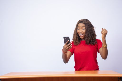 A beautiful young black woman sitting at a desk, looking at her phone and feeling excited