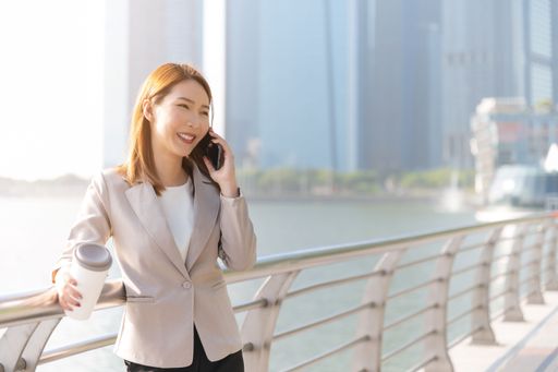 A young Asian businesswoman talking on a phone holding a coffee cup outdoors in a modern city, a business communication concept