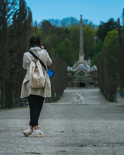 A closeup of a young girl standing on a road surrounded by trees and taking a picture of a building