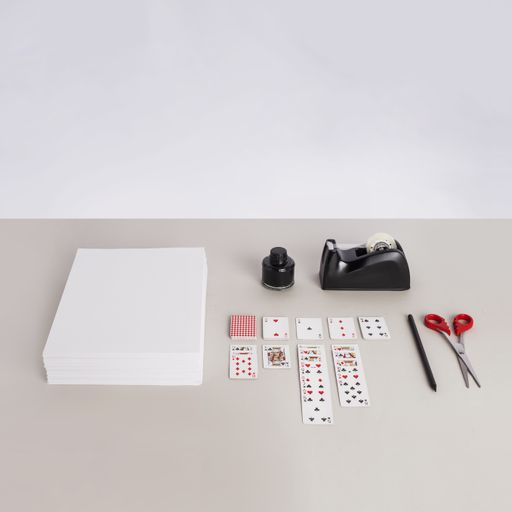White paper, playing cards, scissors, a pencil and an adhesive tape dispenser on a gray surface