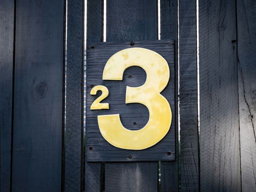 The 2 in 3 house street number sign. Yellow numbers on a black wooden fence.
