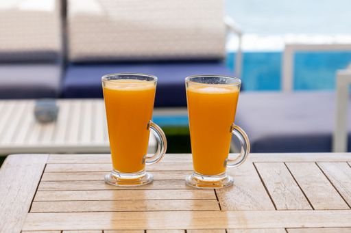 A closeup shot of two glasses of orange juice on a table in a cafe with a blue sky and the sea in its background
