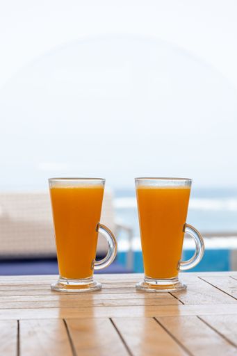A closeup shot of two glasses of orange juice on a table in a cafe with a blue sky and the sea in its background