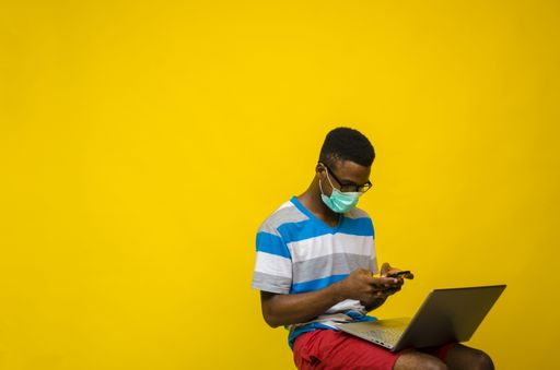 A young black entrepreneur wearing a medical mask and using his smartphone and laptop