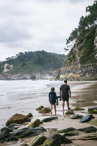 A vertical shot of father and daughter walking on the beach with green-covered hills in front of them