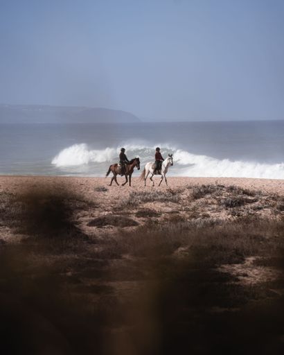 A vertical shot of two men riding horses on the beach in Portugal in daylight