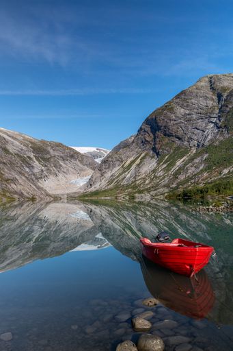 A vertical shot of a rocky mountain with green trees, its reflection in the azure lake, and a red sailboat
