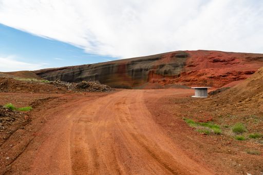 A picturesque rural scene featuring a winding dirt road stretching through a landscape of red dirt hills, leading to an Icelandic open pit mine