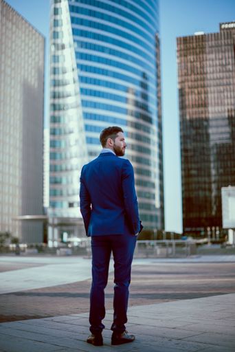 A young businessman in a blue suit standing outside in the downtown, view from the back