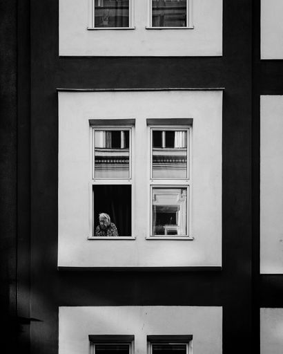 A vertical grayscale shot of an old lady looking out of the window. Vienna, Austria