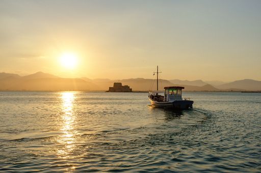 A boat leaves Nafplio harbor and heads toward Bourtzi Castle during sunset