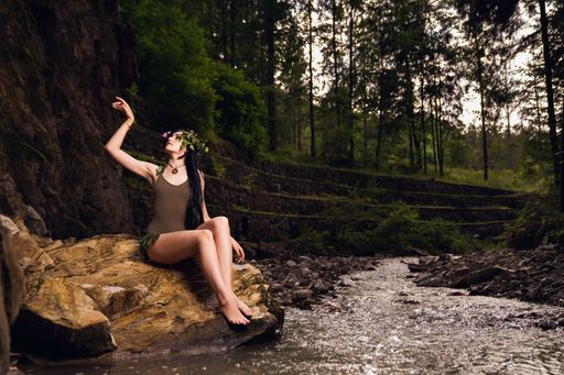 A pretty caucasian female model wearing a flower crown sitting on a rock in the park river