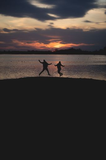 A vertical shot of silhouettes of people jumping up on the shore against dark sunset sky