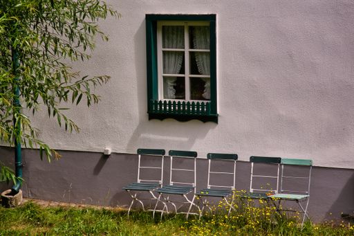 Four old green faded garden chairs in front of a window with grass and flowers growing around them
