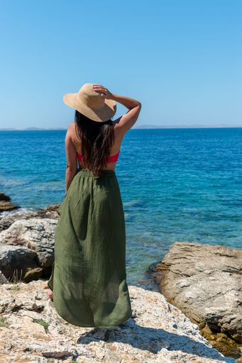 A back view of female wearing summer cloth with hand on head standing on rock in front of sea