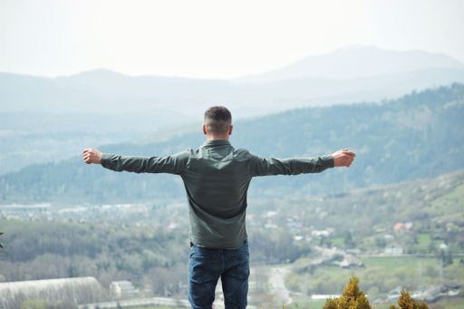 A Caucasian guy standing on top of a hill against green landscape with hands stretched - freedom concept
