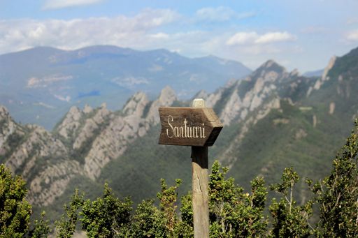 A closeup of a wooden arrow showing the direction to Santuari on the background of forest mountains