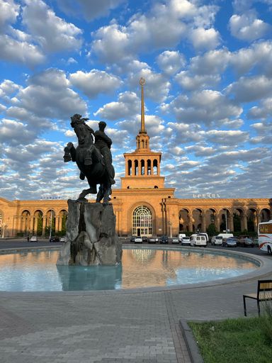 A bronze Sasuntsi David statue in front of a large building subway against a clouded sky
