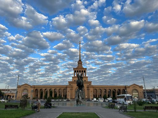 A bronze Sasuntsi David statue in front of a large building subway against a clouded sky