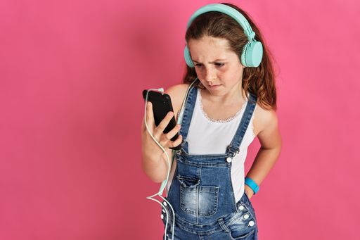 A cute little girl listening to music with a phone and headphones on a pink background