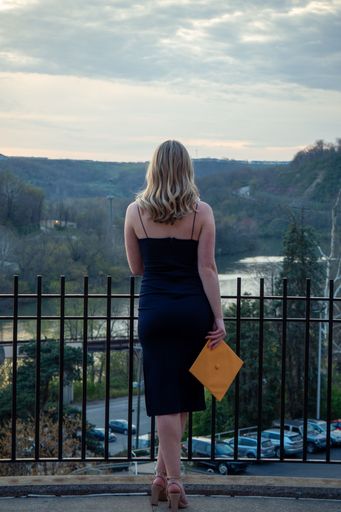 A vertical shot of a girl in a dark blue dress with a yellow mortarboard hat in her hand standing against railings