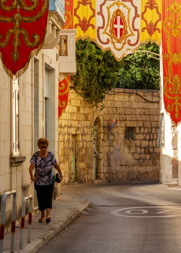 A vertical shot of a lady walking down a narrow street decorated with flags. Lia, Malta.