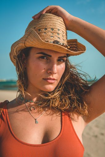 A vertical shot of a European female with an orange swimsuit and a cowboy hat posing on sandy beach