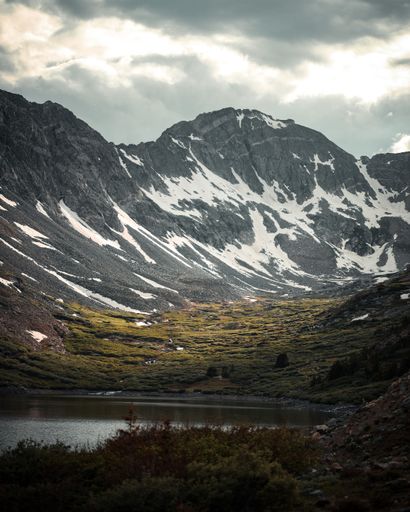 A vertical shot of snowy mountains with clouds and sun rays above and a lake and meadow in the front