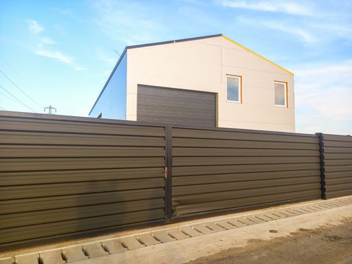 A front view of a fenced warehouse building used as a industrial warehouse, with houses in the background and a clear blue sky
