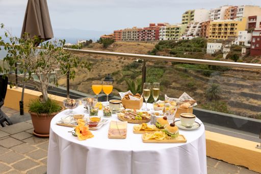 A top closeup of a round table outdoors with a white tablecloth and burgers, chips, two glasses of champagne, white background