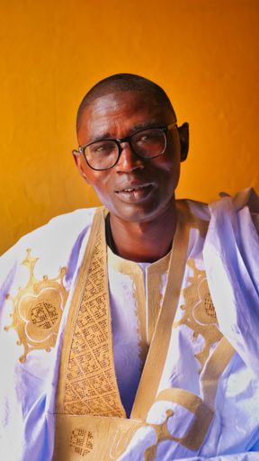 A portrait of an African man against a yellow wall. He wears a typical Mauritanian male dress, the bisht.