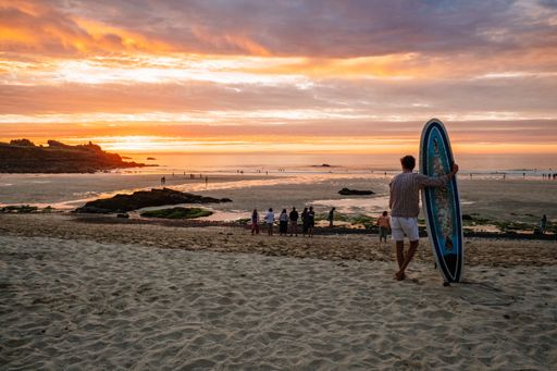 A back shot of an adult man standing on a sandy beach holding surfboard watching the sunset