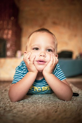 A Caucasian kid resting his chin on hands while lying on his stomach on a cozy carpet, a vertical shot