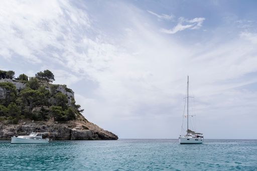a sailboat and yacht, anchored at the entrance of a cove, near a cliff on a summer day with clouds in the sky, cala Galdana, Menorca