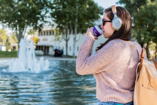 A Caucasian female student wearing wireless headphones and a backpack drinking coffee in a park