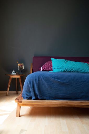 A vertical shot of a cozy, stylish modern bedroom with a blue and red bed beside a small table against the gray wall
